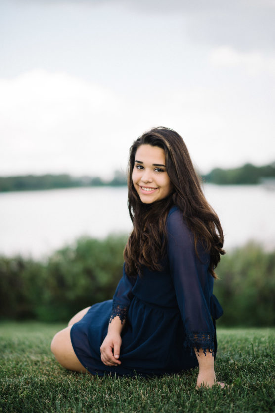 A young woman with long dark hair sits on grass, smiling at the camera. She wears a navy blue dress and has a relaxed, outdoor background with water and greenery behind her. by Justin Salem Meyer
