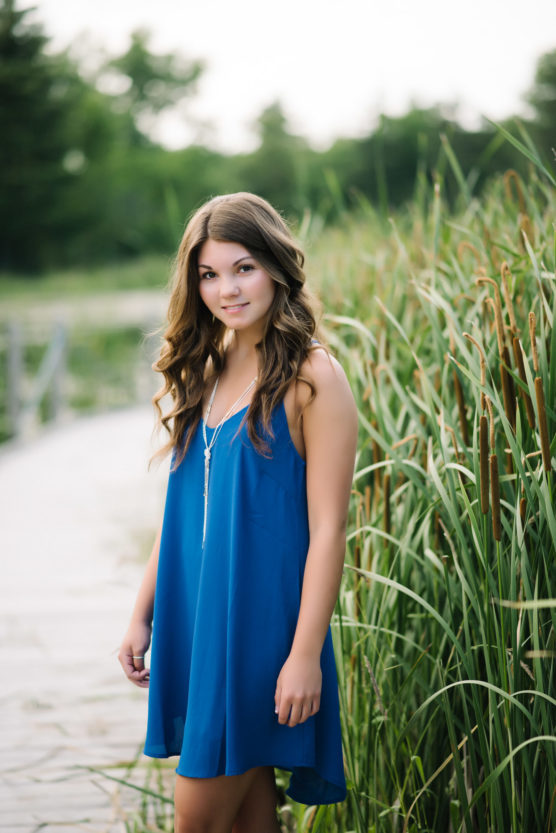 A young woman with long brown hair wearing a sleeveless blue dress stands by tall green grasses outdoors, with a walkway and trees blurred in the background. by Justin Salem Meyer