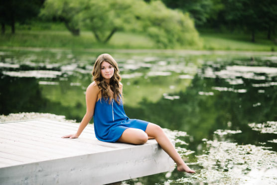 A young woman in a blue dress sits on the edge of a wooden dock, dipping her feet into a calm, green lake surrounded by trees and lush greenery. by Justin Salem Meyer