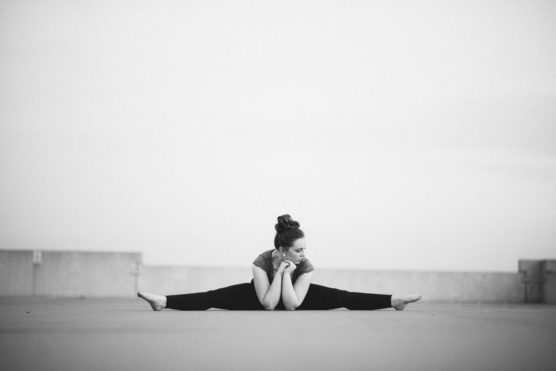 A person sitting on the ground outside, stretching their legs wide apart and leaning forward with arms resting on the floor, looking down pensively. The image is in black and white. by Justin Salem Meyer