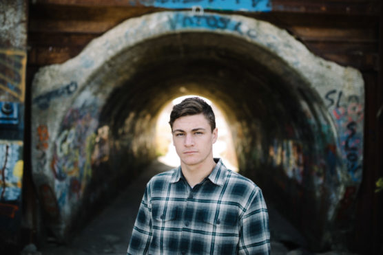 A young man wearing a plaid shirt stands in front of a graffiti-covered tunnel, with sunlight shining through the tunnel behind him. by Justin Salem Meyer