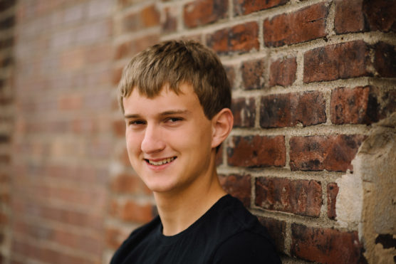 A teenage boy with short light brown hair, wearing a black t-shirt, smiles while leaning against a red brick wall. The background is softly blurred, focusing on his face. by Justin Salem Meyer