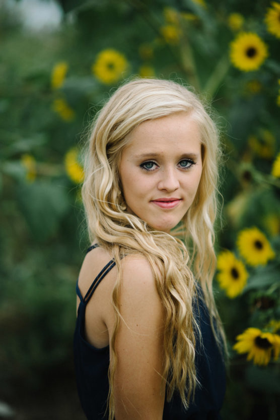 A young woman with long, wavy blonde hair and blue eyes stands outdoors in front of tall green plants with yellow sunflowers, wearing a sleeveless black top and looking over her shoulder at the camera. by Justin Salem Meyer