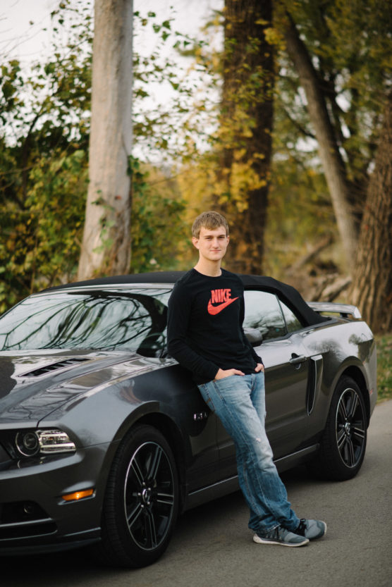 A young man in jeans and a black Nike sweatshirt leans against a gray convertible sports car parked on a tree-lined road, with greenery and tall trees in the background. by Justin Salem Meyer