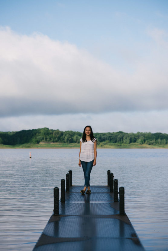 A woman wearing a white top and blue jeans stands and smiles on a dock extending over a calm lake, with green trees and a cloudy sky in the background. by Justin Salem Meyer