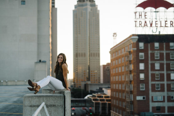 A woman sits on a concrete barrier of a rooftop parking lot, smiling at the camera. Tall buildings and a sign reading “THE TRAVELER” are visible in the background at sunset. by Justin Salem Meyer