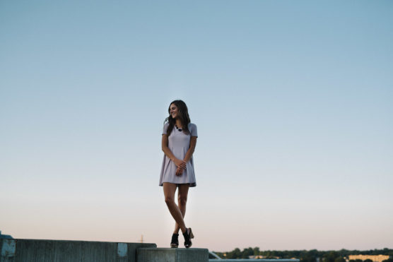A woman in a short-sleeved, light purple dress stands on a concrete ledge outdoors, smiling and looking to the side, with a clear blue sky and trees in the background. by Justin Salem Meyer