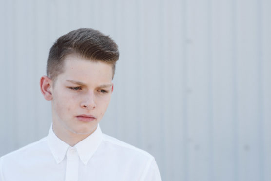 A young person with short brown hair, wearing a white button-up shirt, looks down with a serious expression while standing in front of a light gray, corrugated metal wall. by Justin Salem Meyer
