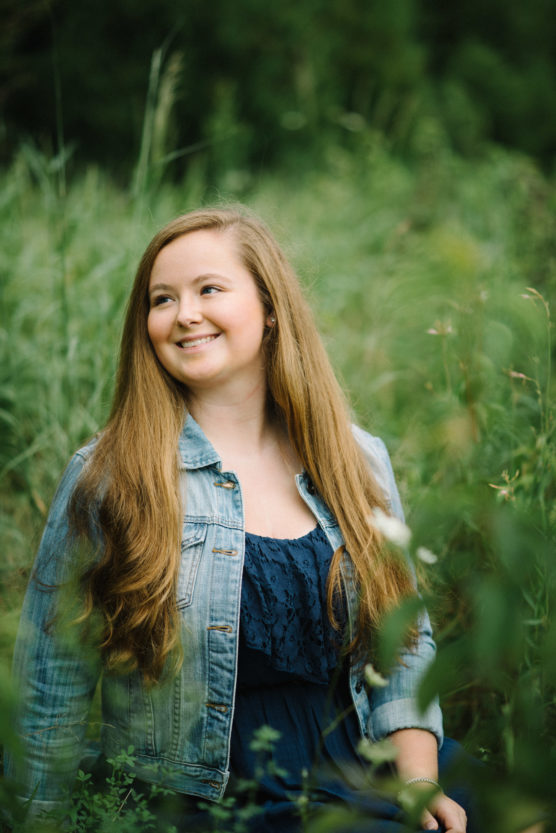 A young woman with long, light brown hair sits in tall green grass, smiling and looking to the side. She is wearing a denim jacket over a dark blue dress. The background is blurred, highlighting her in the natural setting. by Justin Salem Meyer