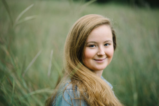 A young woman with long, light brown hair smiles gently while standing in a grassy field, wearing a light blue shirt. The background is softly blurred green foliage. by Justin Salem Meyer