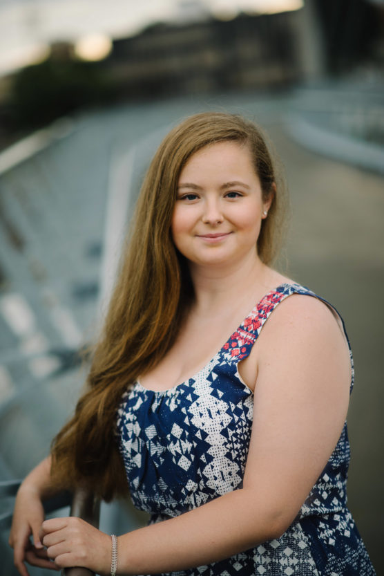 A young woman with long light brown hair wearing a blue and white patterned sleeveless dress stands outdoors by a railing, smiling softly at the camera with a blurred background. by Justin Salem Meyer