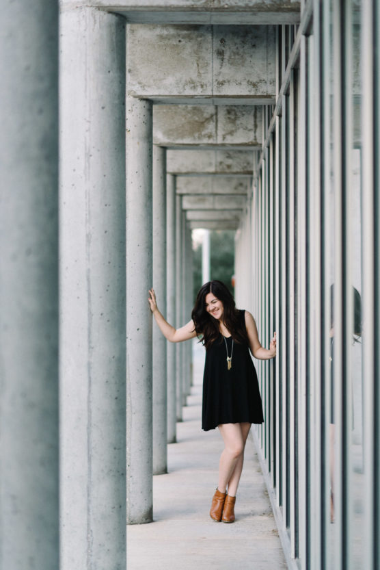 A woman in a black dress and brown ankle boots leans against a row of concrete columns beside a glass wall, smiling with one arm raised and the other touching a column. by Justin Salem Meyer