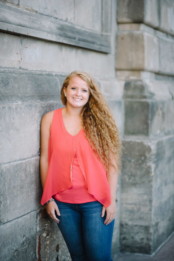 A young woman with long, curly blonde hair wearing a sleeveless coral top and blue jeans stands smiling against a stone wall. by Justin Salem Meyer