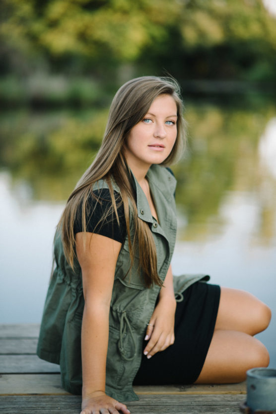 A young woman with long brown hair sits on a wooden dock by a calm lake, wearing a green utility vest over a black dress. The background is blurred with green trees reflected in the water. by Justin Salem Meyer