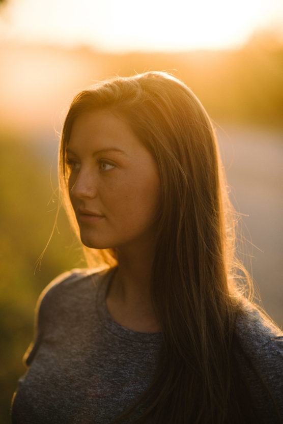 A young woman with long brown hair stands outside at sunset, bathed in warm golden light, looking off to the side. She wears a gray shirt, and the background is softly blurred. by Justin Salem Meyer