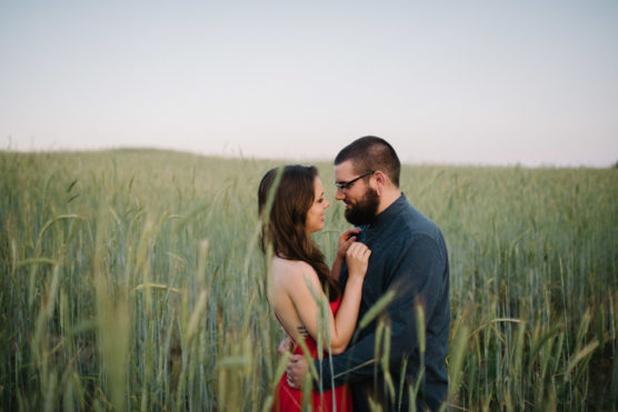 A couple stands close together in a field of tall grass, gazing into each others eyes. The woman wears a red dress and the man wears glasses and a dark shirt. The sky is clear and the mood is intimate. by Justin Salem Meyer
