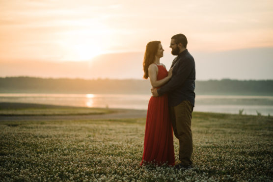 A couple stands facing each other and embracing in a field of flowers by a lake at sunset, with soft, golden light in the background. The woman wears a flowing red dress and the man is dressed in dark pants and a shirt. by Justin Salem Meyer