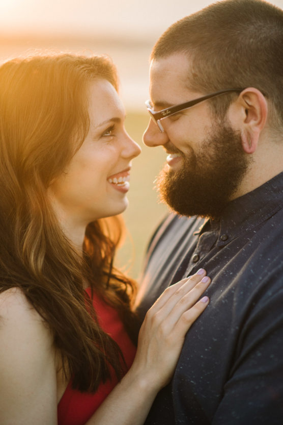 A smiling couple stands close together outdoors at sunset, looking into each other’s eyes. The woman has long brown hair and wears a red top; the man has a beard, glasses, and wears a blue shirt. by Justin Salem Meyer
