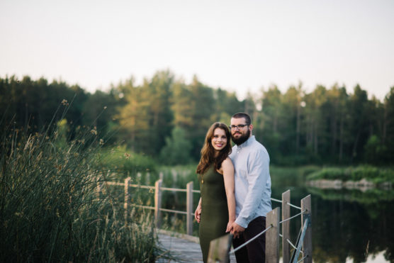 A woman and man stand together on a wooden dock by a lake, holding hands and smiling. Tall grass and trees surround the calm water under a clear sky in the background. by Justin Salem Meyer