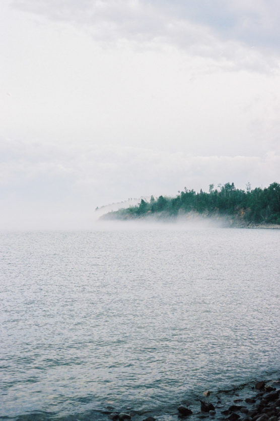 Mist rises from a calm body of water near a rocky shoreline, with a forested peninsula and cloudy sky in the background. by Justin Salem Meyer