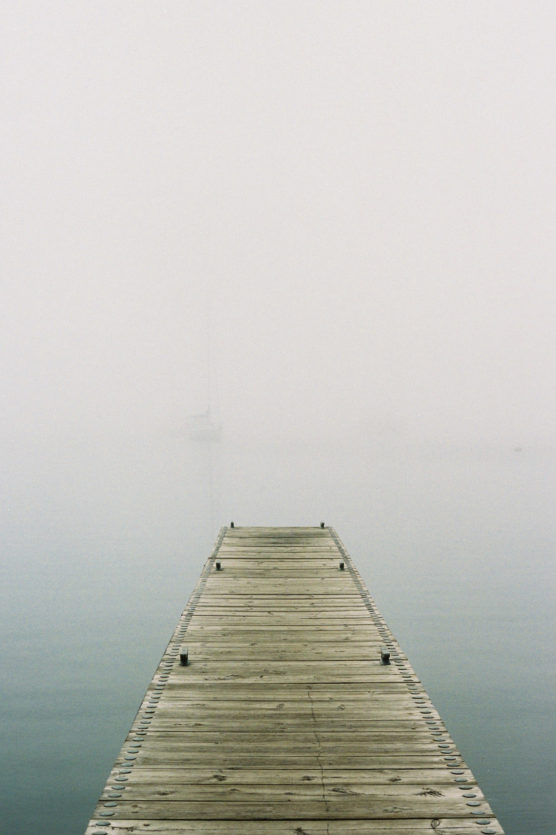 A wooden dock extends over calm water into dense fog, with a faint outline of a sailboat barely visible in the distance. The scene is tranquil and shrouded in mist. by Justin Salem Meyer