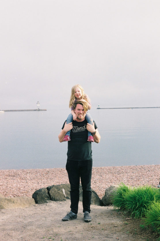 A man stands on a rocky shore by a calm lake, smiling with a young girl sitting on his shoulders. The girl has blonde hair and pink clothes. Theres greenery nearby and a lighthouse visible in the distance. by Justin Salem Meyer