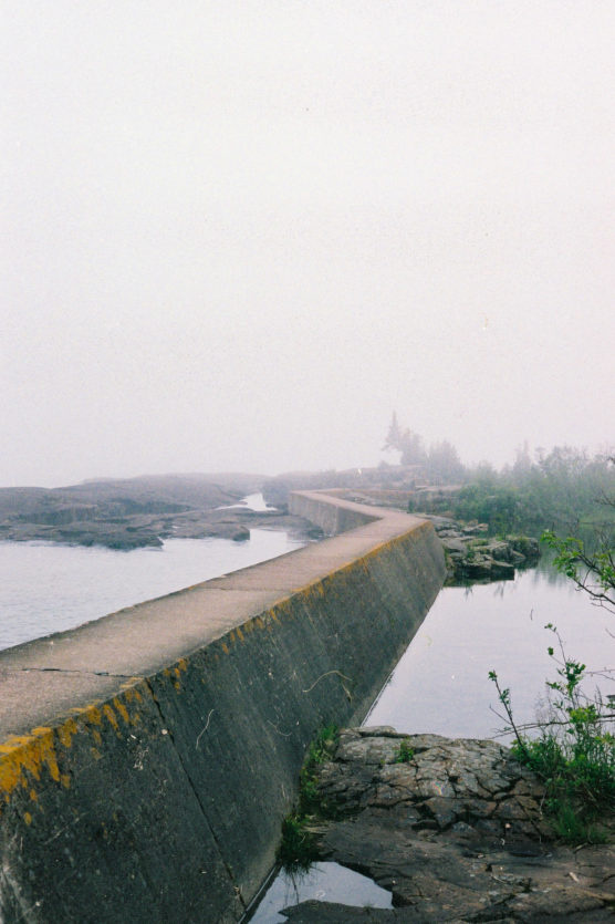 A concrete pier extends into a foggy lake, bordered by rocky shore and green plants. The background fades into mist, obscuring distant trees and water. by Justin Salem Meyer