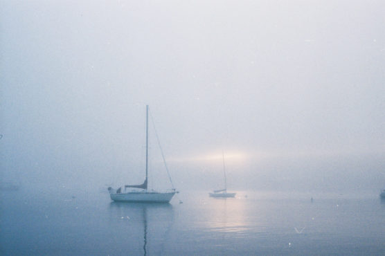 Two sailboats are anchored on calm water in dense fog. The sun is faintly visible through the mist, casting a soft glow and gentle reflections on the water’s surface. The scene looks quiet and serene. by Justin Salem Meyer