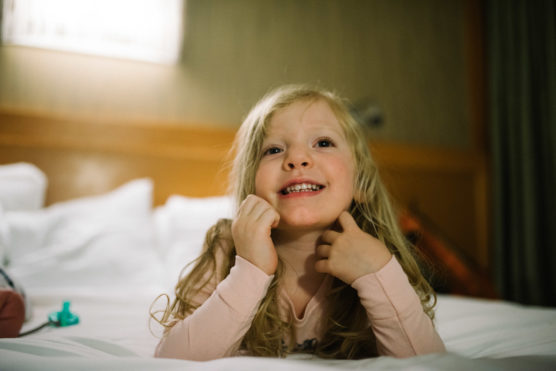 A young girl with long blond hair and a pink shirt lies on a bed, smiling cheerfully and resting her hands on her chin. The background shows a softly lit room with white pillows. by Justin Salem Meyer