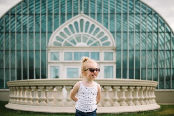 A young child wearing sunglasses and a white polka dot tank top smiles while standing in front of a large, glass-domed conservatory with decorative white columns and geometric window patterns. by Justin Salem Meyer