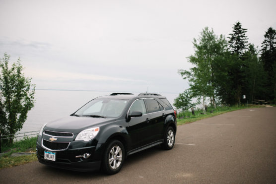 A black Chevrolet SUV is parked in an almost empty lot beside a calm body of water, with trees and greenery along the edge and a cloudy sky above. by Justin Salem Meyer