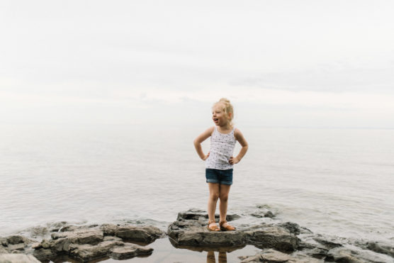 A young girl stands on rocks by the shore, hands on hips, smiling and looking to the side. She wears a white tank top, blue shorts, and sandals. The sky is overcast and the calm water stretches out behind her. by Justin Salem Meyer