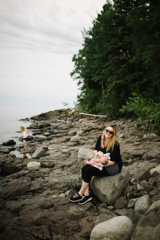 A woman sits on a large rock near a rocky shoreline, holding a baby, while a child plays by the water in the background. Tall trees and overcast sky create a peaceful, natural setting. by Justin Salem Meyer