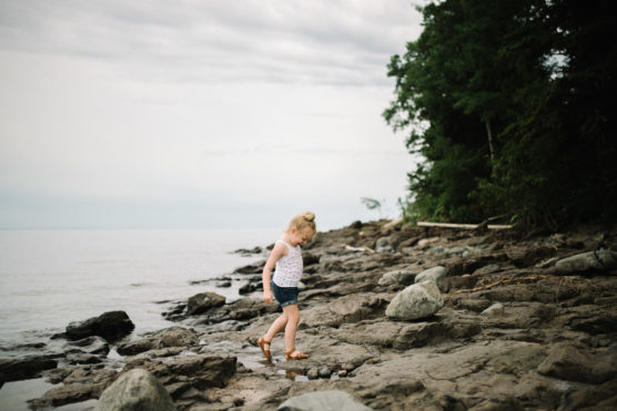 A young girl walks on a rocky shoreline near calm water, with trees on the right and an overcast sky above. She wears a white top, denim shorts, and sandals, looking down at the ground as she walks. by Justin Salem Meyer