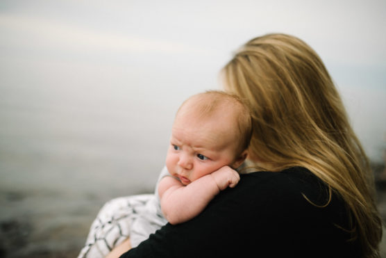 A baby with a curious expression rests on a womans shoulder by the water. The woman has long blonde hair, and both are facing away from the camera towards a calm, hazy shoreline. by Justin Salem Meyer