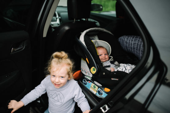 A young child stands smiling outside an open car door, while a baby sits in a car seat inside the car, looking out. The car interior and a patterned pillow are visible. by Justin Salem Meyer