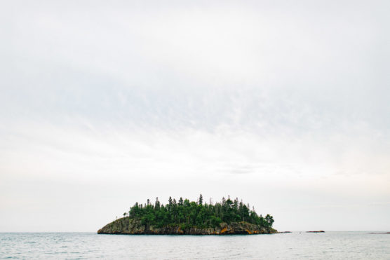 A small, tree-covered island surrounded by calm water under a cloudy sky. The island’s rocky shoreline is visible, and the horizon stretches across the image. by Justin Salem Meyer
