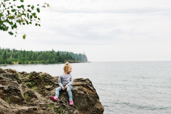 A young child with blond hair, wearing a gray sweatshirt, jeans, and pink shoes, sits on a rocky ledge overlooking a calm lake with a tree-lined shore in the distance under a cloudy sky. by Justin Salem Meyer