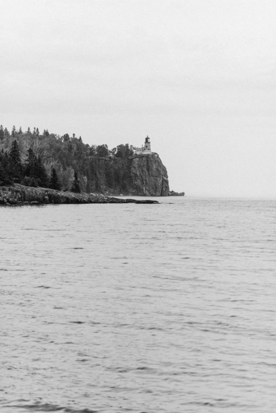 A black-and-white photo showing a rocky shoreline with evergreen trees and a lighthouse perched on a cliff, overlooking a calm, expansive body of water under a cloudy sky. by Justin Salem Meyer