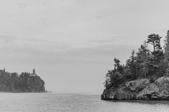Black and white photo of a rocky shoreline with pine trees on the right and a distant lighthouse standing on a cliff to the left, overlooking a calm body of water under a cloudy sky. by Justin Salem Meyer
