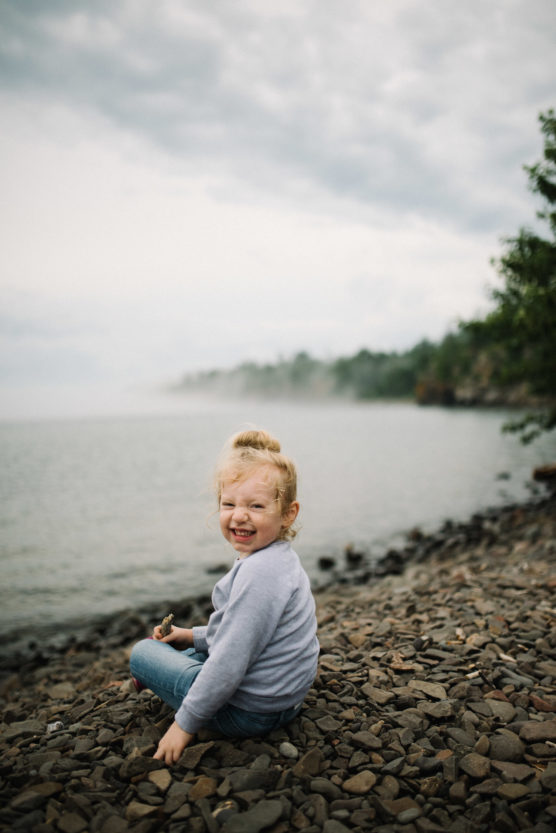 A young child with curly blonde hair sits on a rocky shore, smiling playfully at the camera. The background shows a misty lake and overcast sky, with trees along the shoreline. by Justin Salem Meyer