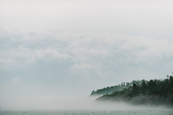 A misty shoreline with a tree-covered cliff partially obscured by fog, meeting calm water under a cloudy sky. by Justin Salem Meyer