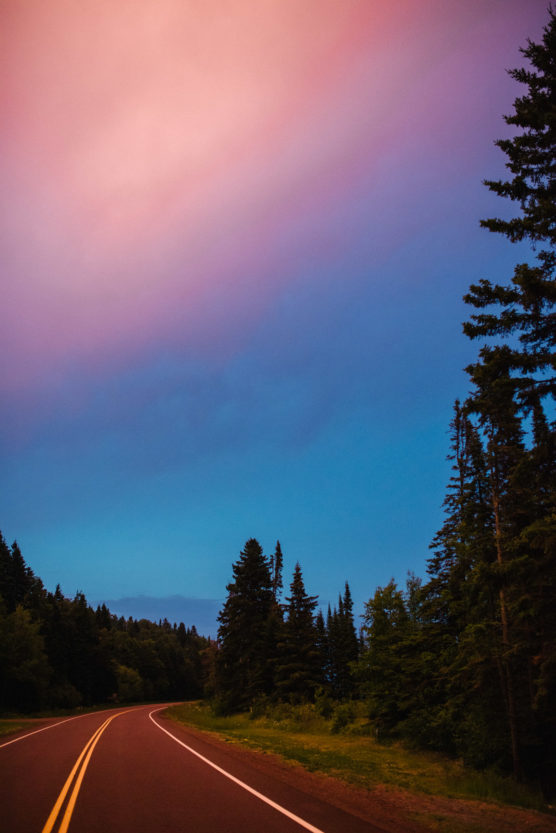 A winding road curves through a forest of tall evergreen trees under a vibrant sky with pink and blue hues at sunset or sunrise. by Justin Salem Meyer