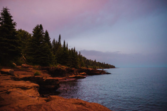 Rocky shoreline bordered by dense evergreen trees under a purple and blue sky at dusk, with calm water stretching to the horizon. by Justin Salem Meyer