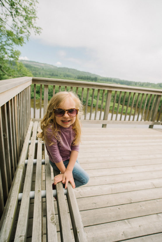 A young girl with long blonde hair, wearing sunglasses, a purple shirt, and jeans, smiles while sitting on a wooden bench on a deck overlooking a scenic, green landscape with trees and hills. by Justin Salem Meyer