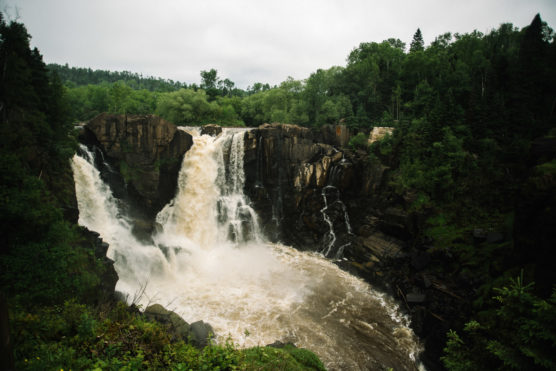 A wide, powerful waterfall cascades over dark rocky cliffs surrounded by dense green forest under an overcast sky. by Justin Salem Meyer
