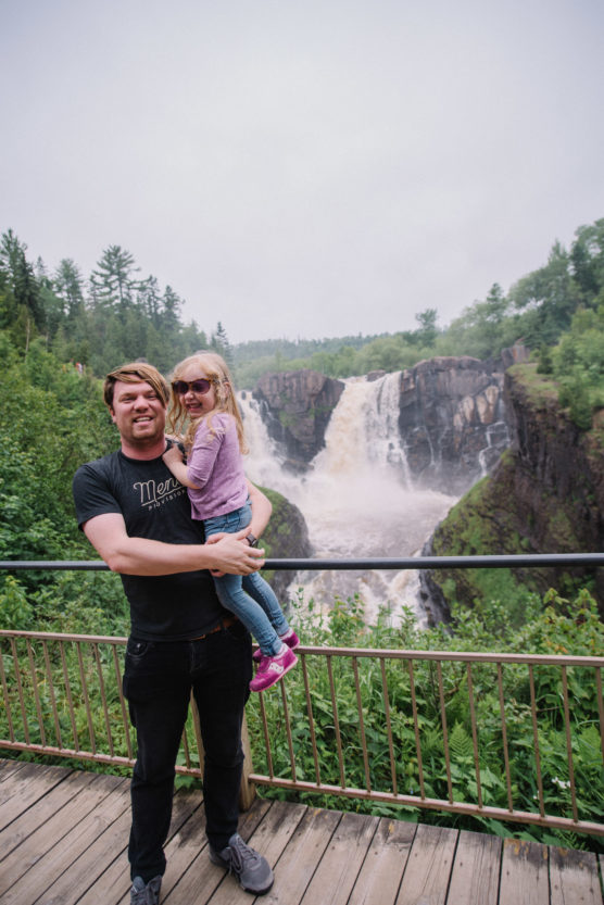 A man holding a young girl stands on a wooden platform in front of a large waterfall surrounded by lush greenery. Both are smiling, and the girl is wearing sunglasses and purple clothes. by Justin Salem Meyer