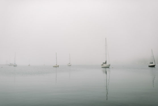 Several sailboats are anchored on calm water under a thick, gray fog, with their reflections visible and the background mostly obscured by mist. by Justin Salem Meyer