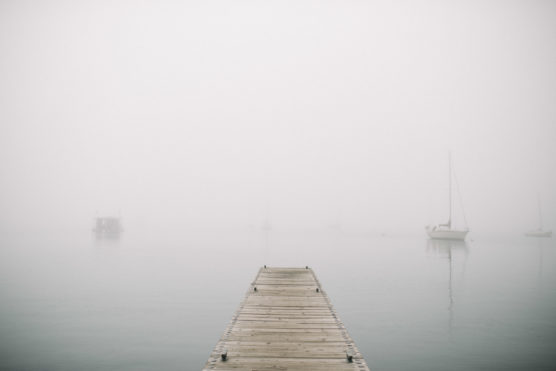 A wooden dock extends over calm water into thick fog, with faint outlines of boats barely visible in the distance. by Justin Salem Meyer