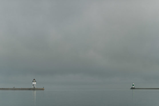 A calm, overcast seascape with two small lighthouses on opposite piers, one white and black on the left and one green and white on the right, against a vast, cloudy sky and smooth water. by Justin Salem Meyer